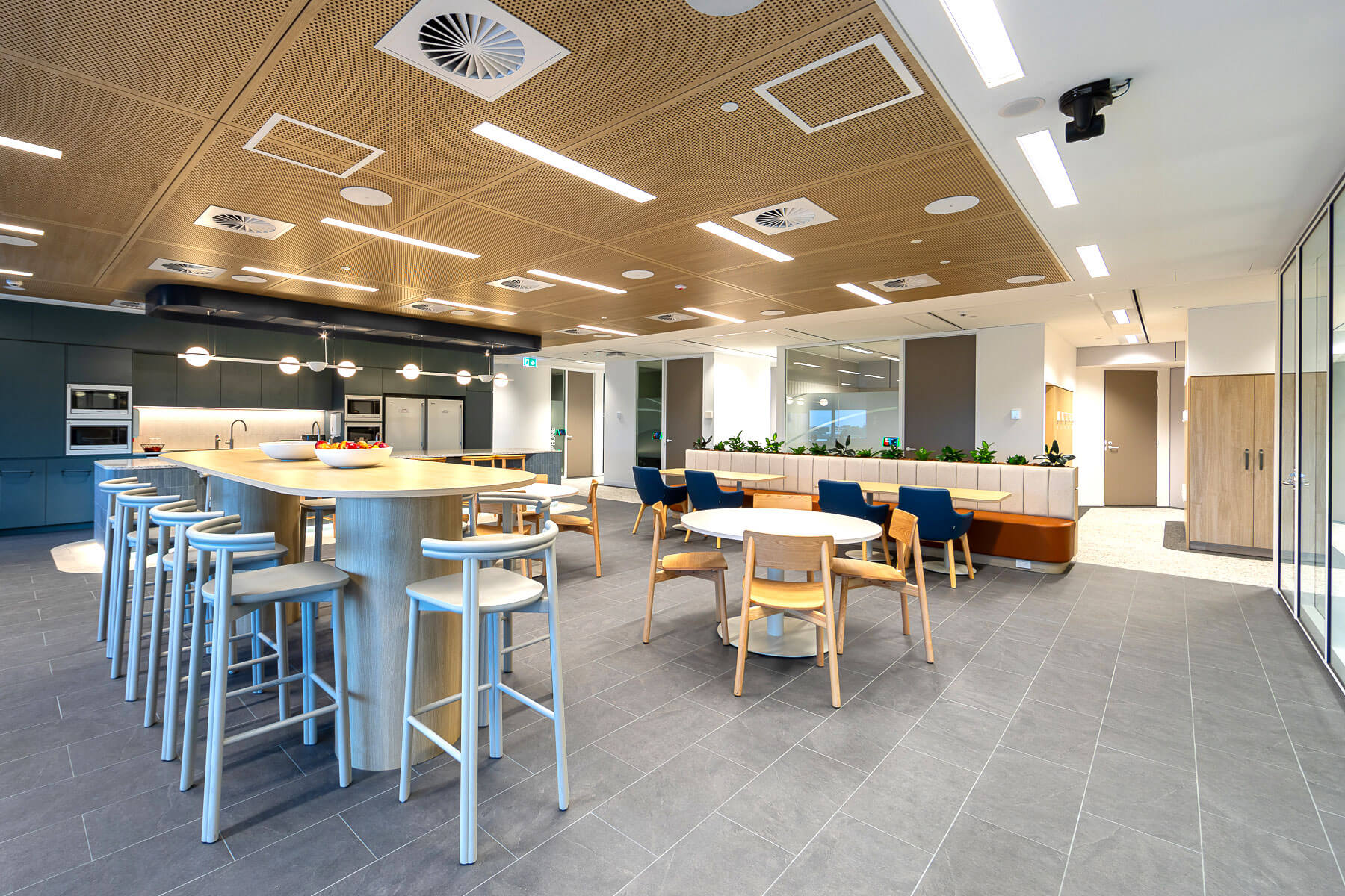 Blue office kitchenette with island bench seating and dining tables behind - alt view