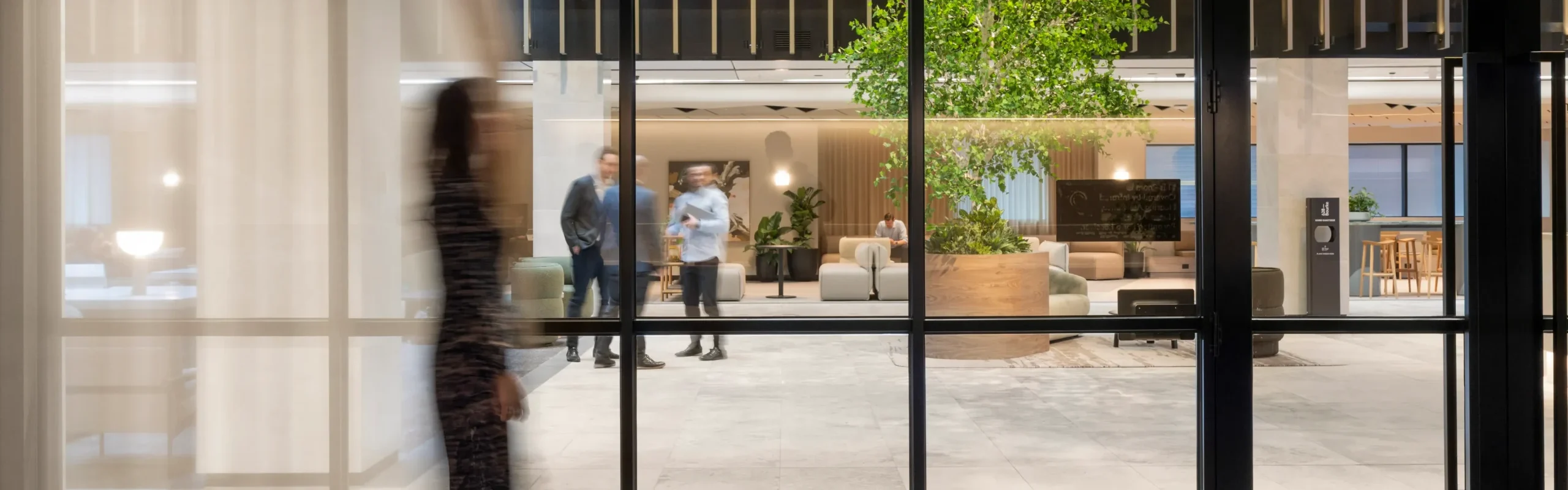View through glass doors into a modern office atrium with a large indoor tree, lounge seating, and people chatting.