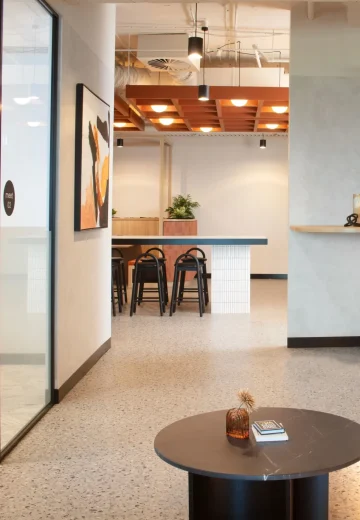 Modern office lounge corner with built-in bench seating, a round coffee table, and a view through to a café-style breakout bar.