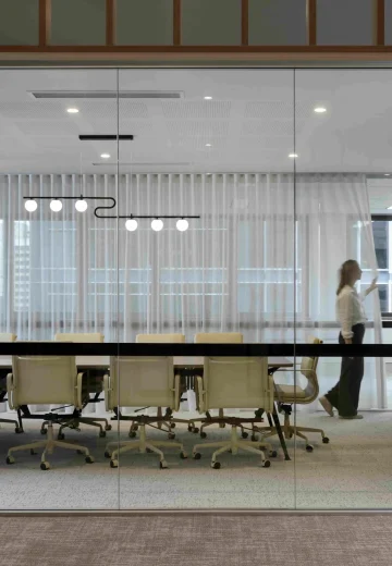 Glass-walled boardroom with a long meeting table, rolling chairs, and sheer curtains, as a person walks past.