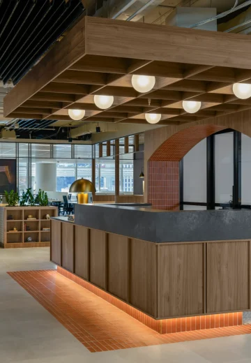 Open-plan office hub with a timber joinery counter, warm underlighting, and a brick-arched feature, leading into work and meeting areas.