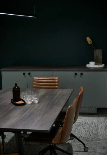 Moody meeting room with a dark wall, timber table, tan leather chairs, and low cabinetry with styled décor.